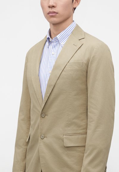 Mand iført beige blazer over en lyseblå og hvidstribet knap-op-skjorte mod en ensfarvet baggrund.