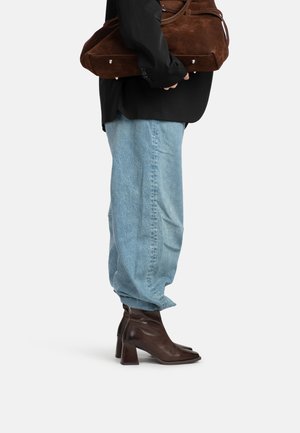 Person trägt hellblaue Jeans und braune Leder-Stiefeletten mit Absatz, hält eine große braune Wildledertasche vor weißem Hintergrund.