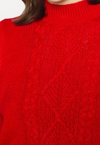 Czerwony sweter z dzianiny z wysokim kołnierzem, z teksturowanymi wzorami w warkocze oraz diamentowym wzorem z przodu. Materiał wydaje się miękki i ciepły.