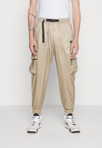 Beige cargo kalhoty vyrobené z lehkého materiálu, s elastickými manžetami, stahovací šňůrkou v pase a dvěma bočními kapsami se zipem.