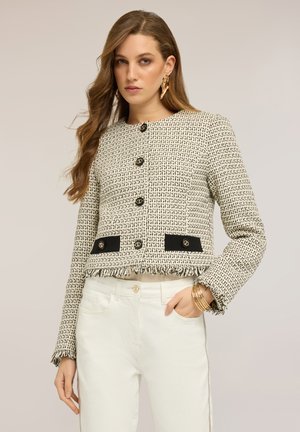 Kvinde med langt bølget hår bærer en tekstureret creme- og sort cropped jakke med guldknapper, hvide bukser, guldringe og armbånd.