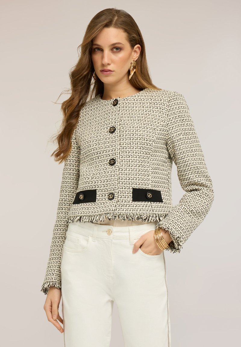 Kvinde med langt bølget hår bærer en tekstureret creme- og sort cropped jakke med guldknapper, hvide bukser, guldringe og armbånd.