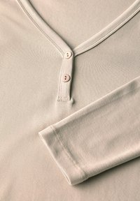 Maglietta beige a maniche lunghe con scollo a V e due piccoli bottoni sul carrè, con la manica destra piegata.