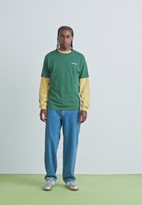 Grøn kortærmet t-shirt lagt over en gul langærmet skjorte, parret med blå straight-leg jeans og grå sneakers.