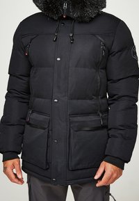Chaqueta negra acolchada con capucha forrada de piel sintética, múltiples bolsillos con cremallera y cierre de botón. Presenta un tejido liso y texturizado.
