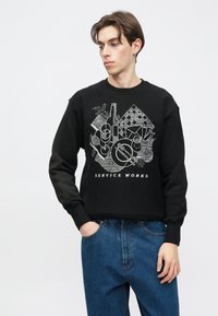 Μαύρο φούτερ με γραφικό σχέδιο διαφόρων αντικειμένων σε λευκό χρώμα, με το κείμενο "SERVICE WORKS" στο κάτω μέρος. Μαλακό, υφή υφάσματος.