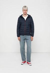 Marineblå pufferjakke med glidelås, hette og røde detaljer. Sammen med lyseblå jeans og røde sneakers. Slett tekstur, quiltet design.