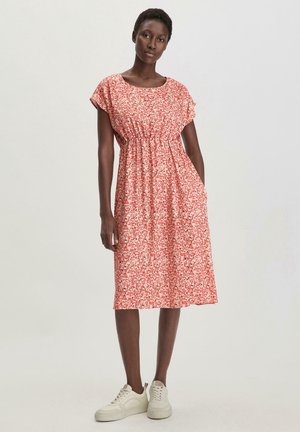 Rotes Blumenkleid mit kurzen Ärmeln, gerafftem Taillenbund, knielangem Saum, aus leichtem Stoff. Kombiniert mit weißen Sneakers.