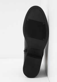 Semelle de chaussure noire avec sections texturées antidérapantes et marquage "Vagabond Shoemakers", taille 37, inclinée contre une surface d’angle blanche.