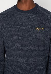 Felpa blu navy realizzata in tessuto testurizzato, con un motivo a pois e colletto a costine. Scritta "Originals" ricamata in giallo sul petto.