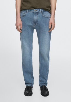 Homme portant un jean droit bleu clair, des mocassins noirs et une chemise vert olive, debout devant un fond blanc uni.