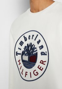 Felpa bianca con un grande logo ricamato circolare con "Timberland" e "Hilfiger" in blu navy e bordeaux. Tessuto liscio, colletto girocollo.