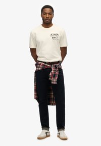 Crèmekleurig katoenen T-shirt met grafische print, donkere jeans en een geruit overhemd om de taille geknoopt. Witte sneakers met lichte accenten.