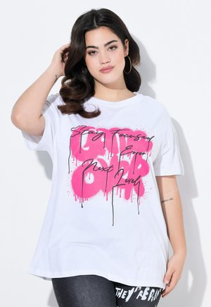 Un t-shirt en coton blanc présente un grand motif rose et noir avec des lettres en coulée. Coupe ample avec des manches courtes, porté avec un jean noir.