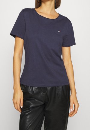 Kobieta w granatowej koszulce z krótkim rękawem z małym logo na piersi oraz czarnych skórzanych spodniach, stojąca z relaksująco opartymi rękami.