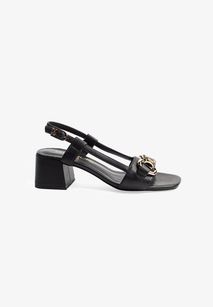 Zwarte leren slingback schoen met een brede blokhak, vierkante tenen en decoratieve gouden hardware detail op de voorste band.
