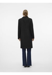 Abrigo negro con un diseño estructurado, mangas largas y un corte recto, combinado con jeans azules acampanados y zapatos de tacón.