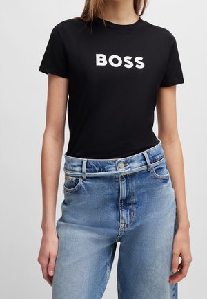 Personne portant un t-shirt noir avec "BOSS" en lettres blanches et un jean taille haute bleu sur un fond uni.
