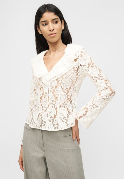 Γυναίκα που φοράει λευκό μακρυμάνικο μπλουζάκι με λουλουδάτη δαντέλα και βολάν στο γιακά, καθώς και γκρι παντελόνι με λεπτές ρίγες, ποζάροντας με το ένα χέρι στη μέση.