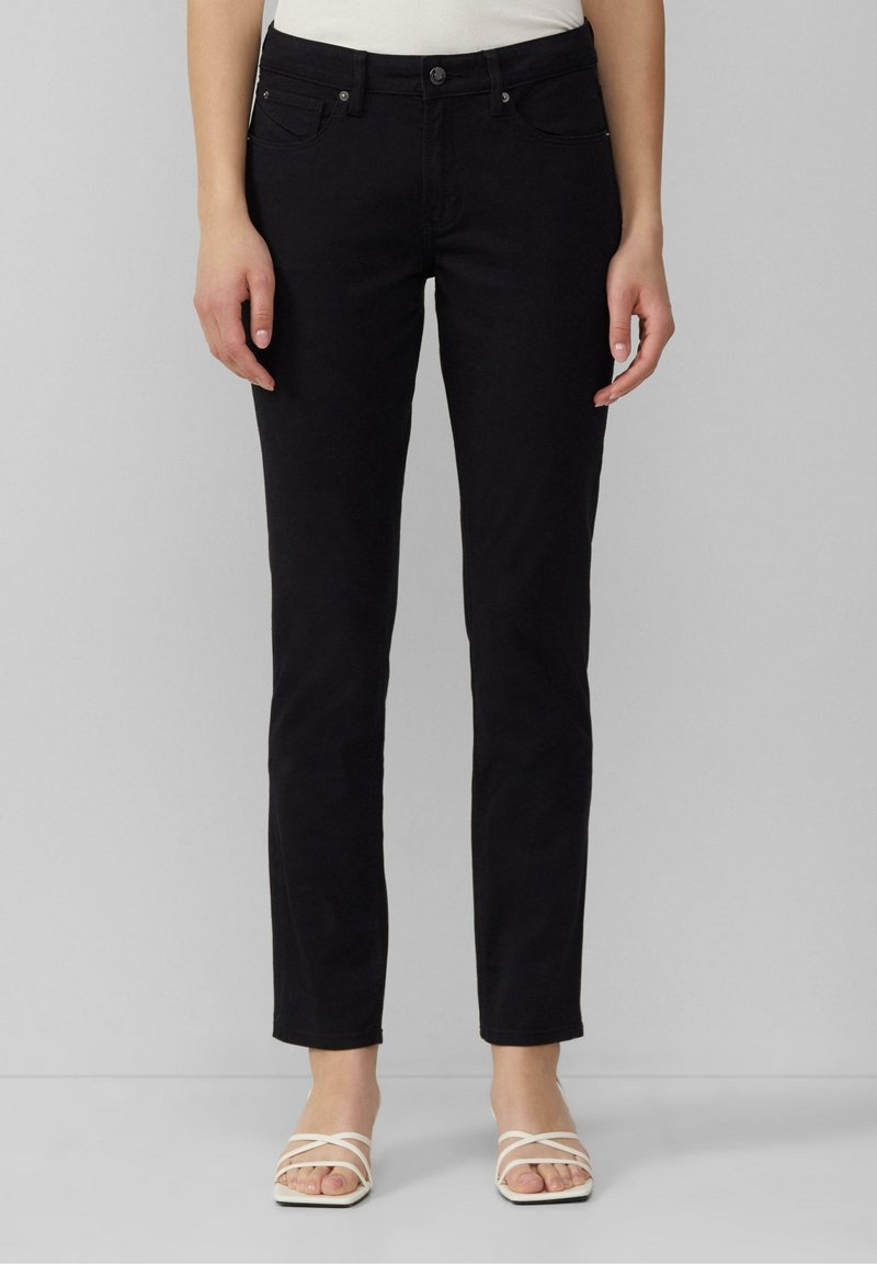 Vue de face d'une personne portant un pantalon noir ajusté et des sandales blanches à lanières, debout devant un fond gris clair uni.