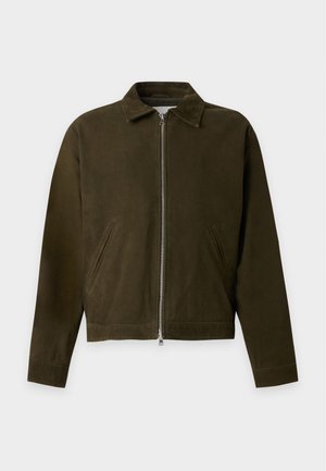 Joven de pie con una chaqueta marrón oscuro con cremallera, pantalones anchos de color gris claro y zapatos marrón oscuro sobre un fondo blanco liso.