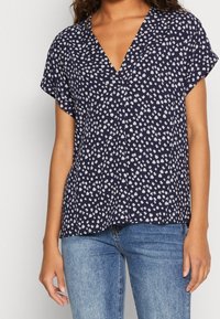 Blusa azul marino con patrón floral blanco, de mangas cortas y escote en V. Fabricación ligera y ajuste holgado, combinada con jeans azules.