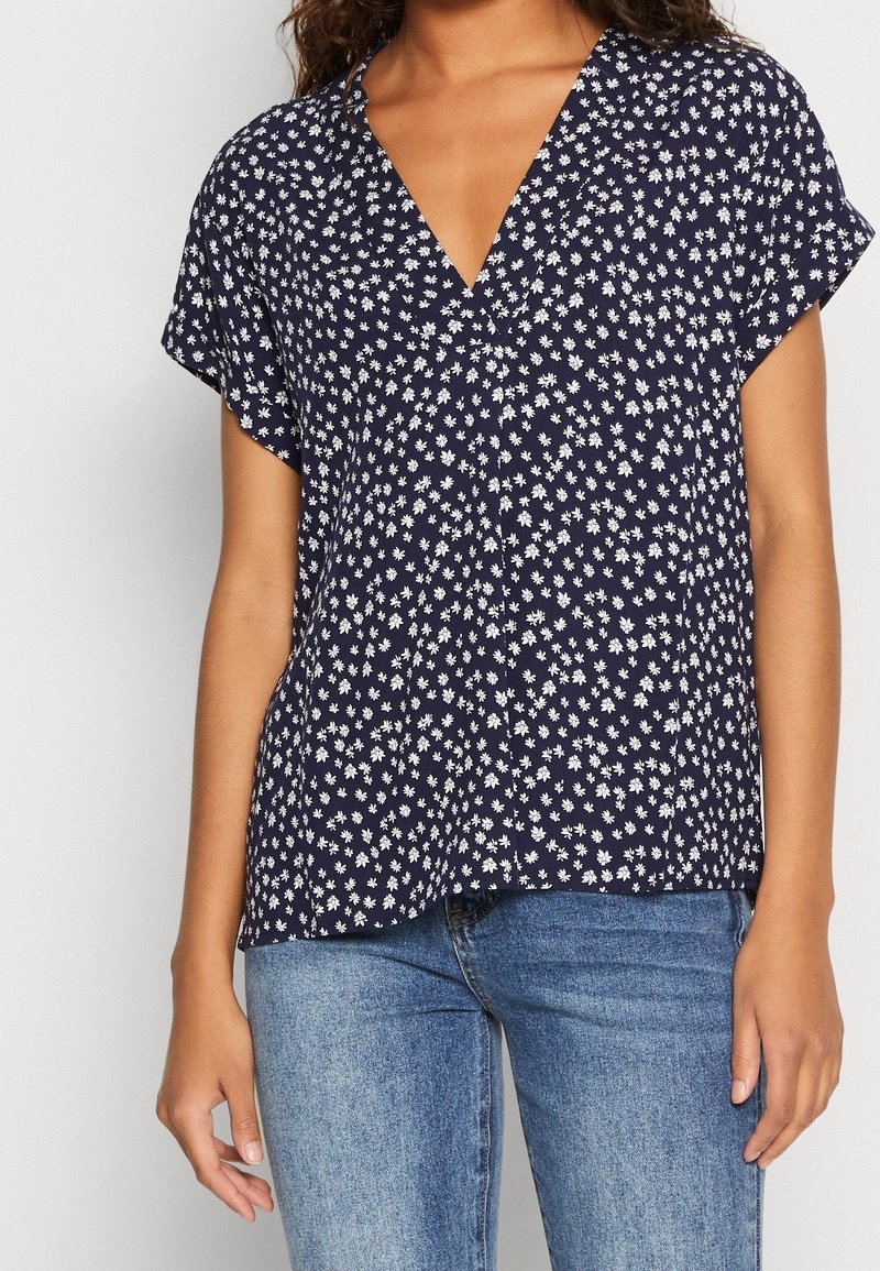 Blusa azul marino con patrón floral blanco, de mangas cortas y escote en V. Fabricación ligera y ajuste holgado, combinada con jeans azules.