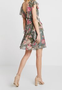 Kobieta w krótkiej, kwiecistej sukience z falbanami, z otwartymi plecami i lekko opadającymi rękawami, w połączeniu z beżowymi, wysokimi sandałami na kostkę z paskiem.