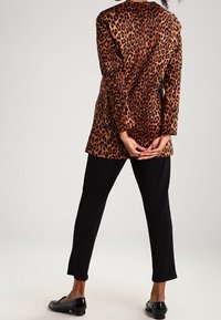 Henkilö seisoo kädet ristissä selän takana, päällään leopardikuosiset pitkät bleiserit, mustat housut ja mustat loafers-kengät.