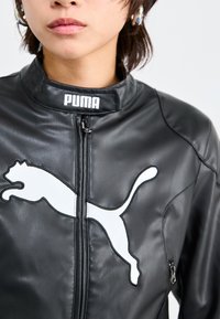 Veste en cuir noir avec un logo Puma blanc surélevé, dotée d'un col montant, d'une fermeture éclair à l'avant et de poches zippées sur les côtés. Texture lisse et coupe ajustée.