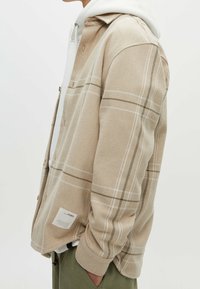 Vue latérale d'une personne portant une veste à boutons beige à carreaux par-dessus un sweat à capuche blanc et un pantalon vert olive, la main dans la poche.