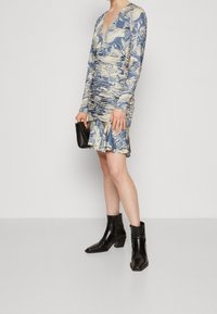 Femme portant une robe florale bleu et beige froncée avec des manches longues, des bottes noires jusqu'aux chevilles, et tenant une petite pochette noire.