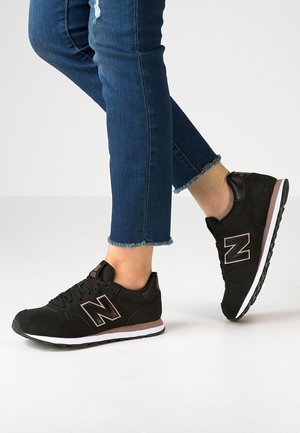 Osoba nosząca czarne i brązowe trampki New Balance z białymi podeszwami oraz niebieskie dżinsy z postrzępionym brzegiem, jedna stopa uniesiona na palcach.