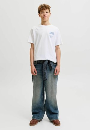Nastolatek o kręconych włosach, ubrany w biały T-shirt z grafiką, niebieską koszulę w kratę zawiązaną w talii, jeansy z szerokimi nogawkami i brązowe buty, stojący na białym tle.