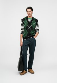 Strickweste mit grünem und schwarzem Diamantmuster über einem gestreiften Schwarz-Weiß-Hemd. Getragen mit dunklen Hosen und braunen Schuhen, hält eine schwarze Tasche.