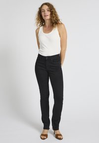 Naine, kes seisab valgel taustal, kandes valget varrukateta toppi, musti kitsaid teksapükse ja pruune kontsakingi sandaale.