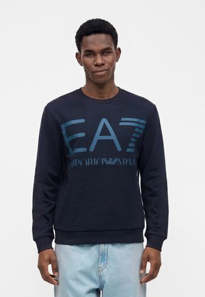 Junger Mann, der einen marineblauen Emporio Armani EA7 Sweatshirt und hellblaue Jeans trägt, vor einem schlichten weißen Hintergrund steht.
