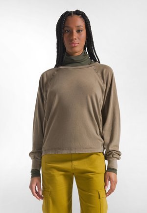 Joven mujer con trenzas largas que lleva un suéter marrón sobre un cuello alto verde y pantalones cargo amarillos mostaza, de pie frente a un fondo blanco.