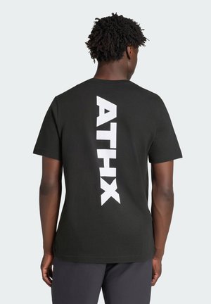 Persona con capelli corti e ricci che indossa una maglietta nera con testo verticale bianco in grassetto "ATHX" sulla schiena, in piedi davanti a uno sfondo uniforme.