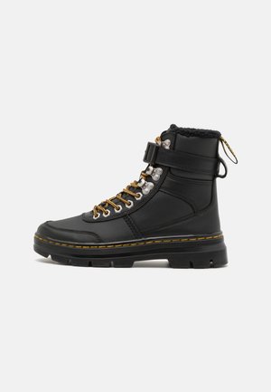 Botte de combat en cuir noir avec lacets jaunes, sangle à la cheville, col matelassé et semelle épaisse et robuste avec détails de couture jaune.