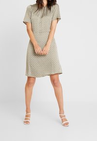 Kleid mit kurzen Ärmeln in einem geometrischen Muster aus Gold, Schwarz und Creme, mit lockerer Passform und knielangem Saum. Dazu werden weiße Riemchensandalen getragen.