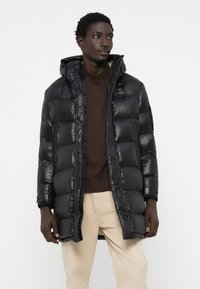 Black puffy jacket with a glossy finish, featuring a hood, zip closure, and multiple block sections. Worn over a dark brown top and beige pants.