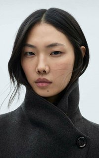 Tmavošedý kabát s vysokým golierom a detailmi na gombíky, vyrobený z mäkkého materiálu. Modelka má stredne dlhé čierne vlasy a prirodzený make-up.