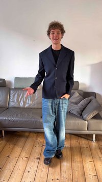 Tengerészkék blézer két első zsebbel, laza világoskék farmernadrággal párosítva. A modell fapadlón áll, egy szürke bőrszofa előtt.