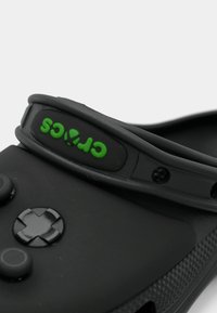 Zwarte Crocs met een groen logo, voorzien van een brede band, zachte gestructureerde materiaal, ronde ventilatiegaten en een rubberen zool.