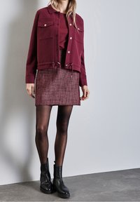 Burgundy cropped jacket with two chest pockets and drawstring hem, paired with a red patterned skirt and black ankle boots.