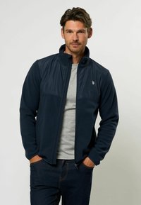 Giacca in pile blu navy con colletto alto e pannello liscio a contrasto. Caratterizzata da zip frontale, tasche laterali e logo ricamato sul petto.