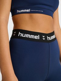 Zbliżenie na osobę noszącą granatowe legginsy sportowe i krótki top z czarnymi elastycznymi paskami z logo "hummel".