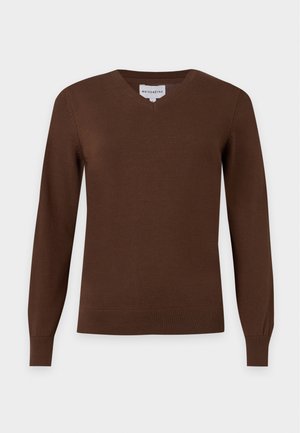 Jeune femme aux longs cheveux blonds portant un pull marron, un pantalon large crème, des chaussures beiges et tenant un sac à main vert olive.