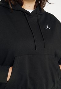 Personne portant un sweat à capuche noir avec un logo Jumpman blanc et les mains dans la poche avant sur un fond uni.